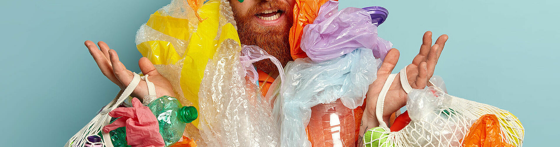 Photo of surprised red haired man has thick beard, overloaded with much garbage, collects plastic, looks with frustrated expression, raises hands, isolated oer blue background. Ecology concept